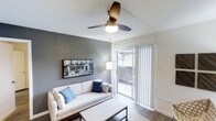 This single-floor residence offers a thoughtfully designed living space with a clean, modern aesthetic. The living room serves as the heart of the home, featuring warm hardwood-style flooring, smooth white ceilings, and a stylish ceiling fan that adds both function and a contemporary touch. An accent wall in a deep charcoal gray creates a bold focal point, complemented by tasteful wall art that enhances the modern atmosphere. Natural light flows generously into the space through large sliding glass doors fitted with vertical blinds, which open directly onto a private outdoor balcony — perfect for enjoying fresh air and outdoor views. The seamless transition between the interior and the balcony gives the living area an open, airy feel. The layout is well-proportioned, comfortably accommodating a sofa and coffee table while maintaining a sense of spaciousness. Overall, the space strikes a balance between comfort and contemporary style, with neutral tones, clean lines, and thoughtful design details that create a welcoming and functional environment.