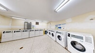 This communal laundry facility offers a practical and efficiently organized space designed to serve multiple users. The room is laid out to maximize usable floor area, with appliances arranged along the perimeter walls to keep the central space open and accessible. A row of front-loading washing machines lines one wall, while top-loading dryers are positioned along an adjacent wall, providing a comprehensive laundering setup. Exposed utility piping is visible in one corner, underscoring the room's functional character. A freestanding utility sink is conveniently located near the entrance, offering an additional washing station for hand-washing or pre-treating garments. Informational signage is posted on the walls, providing usage instructions and guidelines for residents. The flooring throughout is durable terrazzo tile, well-suited for a high-traffic utility environment. Overhead fluorescent tube lighting ensures the space is brightly and evenly illuminated. A baseboard heater along one wall provides climate comfort, and a fire extinguisher is mounted near the exit for safety. Two access doors offer entry and exit points, contributing to smooth traffic flow. The pale yellow-toned walls and white appliances give the room a clean, utilitarian aesthetic.