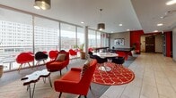 Located at 200 W Norwich Ave in Columbus, Ohio, this dynamic single-floor amenity space is designed to foster community and recreation. The interior is defined by a bold red and gray color palette, lending it an energetic, modern character throughout.

The central lounge area is a standout feature, boasting floor-to-ceiling glass windows that flood the space with natural light and offer sweeping views of the Columbus skyline. Comfortable red mid-century-style lounge chairs and sofas are arranged around a striking red patterned area rug, creating an inviting gathering spot. A wall-mounted flat-screen TV set against a vivid red accent wall anchors a dedicated media and seating nook, complemented by additional lounge chairs and tropical potted plants.

A full-size ping pong table occupies the game zone, set on carpet flooring alongside a corridor that leads deeper into the space. Adjacent to the game area, a kitchenette features a breakfast bar lined with bold red metal stools, pendant lighting, white cabinetry, and a pass-through window. Additional rooms visible through glass-panel doors suggest space for private meetings or study.

The entry area is finished with light tile flooring and features clearly marked access doors with red accent columns. Recessed lighting throughout ensures consistent, well-distributed illumination across all areas of the space.