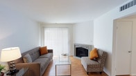 This single-floor space offers a clean, modern aesthetic defined by crisp white walls and warm hardwood flooring that runs throughout. The living area serves as the focal point of the space, anchored by an elegantly detailed fireplace with a decorative mantle that adds a classic touch to the otherwise contemporary setting. Natural light floods the room through a large sliding glass door dressed with full-length vertical blinds, creating a bright and airy atmosphere. The living area is thoughtfully arranged with a tufted gray sofa and a patterned accent chair, both accented with warm orange throw pillows that add a pop of color against the neutral palette. A sleek glass-top coffee table rests at the center, contributing to the space's light and open feel. The white ceilings enhance the sense of openness, while the overall design strikes a balance between comfort and simplicity. The combination of natural light, warm flooring tones, and the inviting fireplace creates a welcoming ambiance well-suited for relaxed everyday living.