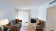 This single-floor space offers a clean, modern aesthetic defined by crisp white walls and warm hardwood flooring that runs throughout. The living area serves as the focal point of the space, anchored by an elegantly detailed fireplace with a decorative mantle that adds a classic touch to the otherwise contemporary setting. Natural light floods the room through a large sliding glass door dressed with full-length vertical blinds, creating a bright and airy atmosphere. The living area is thoughtfully arranged with a tufted gray sofa and a patterned accent chair, both accented with warm orange throw pillows that add a pop of color against the neutral palette. A sleek glass-top coffee table rests at the center, contributing to the space's light and open feel. The white ceilings enhance the sense of openness, while the overall design strikes a balance between comfort and simplicity. The combination of natural light, warm flooring tones, and the inviting fireplace creates a welcoming ambiance well-suited for relaxed everyday living.