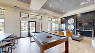 Located at 7430 N 100th Ave in Glendale, Arizona, this single-floor community space offers a thoughtfully designed, open-concept layout that seamlessly blends social and leisure areas. The interior is anchored by soaring ceilings with a striking geometric patterned ceiling feature that adds architectural drama to the main gathering area. Light wood-look tile flooring runs throughout, lending a warm and cohesive feel to the space.

The kitchen and living area is outfitted with sleek dark cabinetry, expansive white countertops, open shelving, a herringbone tile backsplash, and a modern globe pendant chandelier. Large sliding glass doors and clerestory windows flood the space with natural light and provide direct views of the outdoor courtyard.

The main lounge area features a full-size billiards table as a central focal point, surrounded by contemporary curved seating, accent chairs, and layered area rugs in warm gold and neutral tones. A dark shiplap accent wall with built-in display shelving and circular mirrors creates a sophisticated backdrop.

An adjacent lounge wing offers flexible seating with tufted benches, leather swivel chairs, built-in desk stations, and wall-mounted displays — ideal for both relaxation and productivity. Curated artwork and intentional lighting complete the polished, modern aesthetic throughout.