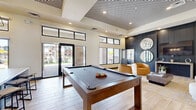 Located at 7430 N 100th Ave in Glendale, Arizona, this single-floor community space offers a thoughtfully designed, open-concept layout that seamlessly blends social and leisure areas. The interior is anchored by soaring ceilings with a striking geometric patterned ceiling feature that adds architectural drama to the main gathering area. Light wood-look tile flooring runs throughout, lending a warm and cohesive feel to the space.

The kitchen and living area is outfitted with sleek dark cabinetry, expansive white countertops, open shelving, a herringbone tile backsplash, and a modern globe pendant chandelier. Large sliding glass doors and clerestory windows flood the space with natural light and provide direct views of the outdoor courtyard.

The main lounge area features a full-size billiards table as a central focal point, surrounded by contemporary curved seating, accent chairs, and layered area rugs in warm gold and neutral tones. A dark shiplap accent wall with built-in display shelving and circular mirrors creates a sophisticated backdrop.

An adjacent lounge wing offers flexible seating with tufted benches, leather swivel chairs, built-in desk stations, and wall-mounted displays — ideal for both relaxation and productivity. Curated artwork and intentional lighting complete the polished, modern aesthetic throughout.