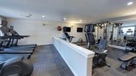 This fitness center offers a practical and organized workout environment designed to support a range of exercise routines. The space is finished with neutral white walls and recessed ceiling lighting that creates a bright, evenly lit atmosphere conducive to focused training sessions. Patterned carpet tiles in muted blue and gray tones cover the floor, adding visual texture while providing a comfortable surface underfoot.

The facility is divided into distinct workout zones. A row of cardio machines, including stationary bikes and upright cycling equipment, lines one side of the space, separated by a low white partition wall that helps define the layout. Multiple wall-mounted flat-screen televisions are positioned for easy viewing from the cardio area, enhancing the workout experience. On the opposite side, a multi-function cable strength training machine offers resistance-based exercise options. Exercise balls are also available, adding versatility for core and balance training. The overall design is clean and straightforward, prioritizing functionality and ease of use for a well-rounded fitness experience.