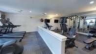 This fitness center offers a practical and organized workout environment designed to support a range of exercise routines. The space is finished with neutral white walls and recessed ceiling lighting that creates a bright, evenly lit atmosphere conducive to focused training sessions. Patterned carpet tiles in muted blue and gray tones cover the floor, adding visual texture while providing a comfortable surface underfoot.

The facility is divided into distinct workout zones. A row of cardio machines, including stationary bikes and upright cycling equipment, lines one side of the space, separated by a low white partition wall that helps define the layout. Multiple wall-mounted flat-screen televisions are positioned for easy viewing from the cardio area, enhancing the workout experience. On the opposite side, a multi-function cable strength training machine offers resistance-based exercise options. Exercise balls are also available, adding versatility for core and balance training. The overall design is clean and straightforward, prioritizing functionality and ease of use for a well-rounded fitness experience.