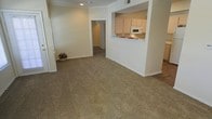 This single-story space features an open-concept layout that promotes a sense of connectivity between the living area and kitchen. The living room is finished with wall-to-wall carpeting in a neutral, earthy tone, providing a comfortable and cohesive foundation throughout the space. Smooth, light-colored walls with white baseboards contribute to a clean and understated aesthetic.

The kitchen is positioned to the right and is accessible via a pass-through counter that opens directly to the living area, creating an integrated feel ideal for everyday living. White cabinetry lines the upper kitchen walls, complemented by a built-in white microwave. A tiled countertop with a small decorative accent adds a subtle touch of color to the otherwise neutral palette.

A doorway visible from the main living area leads down a hallway to additional rooms, suggesting a functional layout beyond what is immediately visible. Recessed lighting in the ceiling provides focused illumination, while the overall design emphasizes practicality and simplicity. The neutral color scheme throughout makes the space adaptable to a variety of interior styles.
