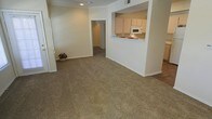 This single-story space features an open-concept layout that promotes a sense of connectivity between the living area and kitchen. The living room is finished with wall-to-wall carpeting in a neutral, earthy tone, providing a comfortable and cohesive foundation throughout the space. Smooth, light-colored walls with white baseboards contribute to a clean and understated aesthetic.

The kitchen is positioned to the right and is accessible via a pass-through counter that opens directly to the living area, creating an integrated feel ideal for everyday living. White cabinetry lines the upper kitchen walls, complemented by a built-in white microwave. A tiled countertop with a small decorative accent adds a subtle touch of color to the otherwise neutral palette.

A doorway visible from the main living area leads down a hallway to additional rooms, suggesting a functional layout beyond what is immediately visible. Recessed lighting in the ceiling provides focused illumination, while the overall design emphasizes practicality and simplicity. The neutral color scheme throughout makes the space adaptable to a variety of interior styles.