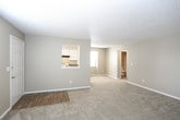 This single-floor residential unit is thoughtfully laid out to maximize functionality and comfort throughout. The living area serves as the heart of the home, featuring plush wall-to-wall carpeting, warm beige walls, and a wall-mounted HVAC unit positioned beneath a window fitted with blinds that allow for natural light control. A pass-through opening connects the living space to the kitchen, creating an open, communicative flow between rooms.

The kitchen is equipped with a full suite of appliances, including a black refrigerator and a white electric coil range with an overhead exhaust hood. White cabinetry lines the upper walls, providing ample storage, while durable laminate wood flooring adds a practical and clean finish underfoot. A window brightens the kitchen with natural light.

The bedroom is generously sized with soft carpeting and a sliding glass door fitted with vertical blinds, offering direct access to the outdoors and a view of the surrounding greenery. A baseboard heater runs along the exterior wall. The bedroom connects to a spacious walk-in closet outfitted with built-in shelving and a hanging rod, providing extensive storage options.

The bathroom features a clean, white soaking tub and shower combination with chrome fixtures and a towel bar. Textured ceilings are found throughout the unit, and white baseboards provide crisp contrast against the neutral wall palette, lending a tidy, well-maintained ambiance across all spaces.