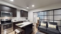 This single-floor studio is thoughtfully designed with a contemporary open-concept layout that maximizes functionality and style. The kitchen is a standout feature, showcasing rich dark espresso cabinetry paired with light marble-style countertops that extend into a peninsula with bar seating — ideal for casual dining or entertaining. The kitchen is equipped with recessed lighting overhead, a stainless steel sink, and dedicated counter space for appliances. Light hardwood-style flooring runs throughout, tying the kitchen and living areas together seamlessly. The living area is intimate yet well-appointed, centered around a leather accent chair with decorative pillows in a complementary yellow and grey palette. A rustic wooden side table and a soft area rug add warmth and texture to the space. A striking black-framed glass partition wall serves as a bold architectural divider, adding an industrial-modern element while allowing light to filter through. A separate laundry area with stacked units is visible beyond the partition. Recessed ceiling lights provide consistent, modern illumination throughout. The overall ambiance is sleek and cohesive, blending dark and light contrasts with thoughtful design details suited for efficient, stylish living.