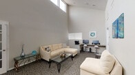 This single-floor space functions as a combined lounge and business center, distinguished by its dramatically vaulted ceilings that lend an open, airy quality to the interior. The soaring ceiling height is accentuated by a row of clerestory windows positioned near the roofline, allowing natural light to filter in from above and illuminate the neutral gray walls throughout.

The lounge area is furnished with a cream sectional sofa and a coordinating loveseat, arranged around a glass-top coffee table with a geometric metal base. Decorative accents, including patterned throw pillows and small sculptural objects, add visual interest to the seating zone. A delicate glass side table with ornate metalwork provides additional surface space.

Along one wall, a built-in granite-topped workstation serves as a dedicated business center, equipped with two computer monitors, a printer, and bar-height seating on chrome stools. Wall-mounted artwork in teal tones complements the overall color palette.

The space features textured, patterned carpet throughout, contributing warmth underfoot. A glass-paned French door offers a refined architectural detail and connects to an adjacent area, while standard entry doors and slatted window blinds complete the functional design. The overall ambiance is clean, modern, and purposefully versatile.