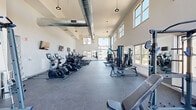 Located at 266 Morris Ave in Long Branch, NJ, this well-equipped fitness center offers a comprehensive exercise facility across a single, expansive floor. The space is defined by its striking architectural design, featuring soaring double-height ceilings with exposed industrial-style ductwork and pendant lighting that add a modern, urban character to the room.

Large floor-to-ceiling windows line the walls, flooding the interior with abundant natural light and offering views of the surrounding neighborhood. The open layout is maximized for functionality, with durable rubber flooring throughout providing both safety and comfort underfoot.

The cardio zone is well-stocked with multiple treadmills, elliptical trainers, stationary bikes, and rowing machines, all arranged to take advantage of the natural light and exterior views. The strength training area includes a multi-station cable machine, a power rack with an Olympic barbell and weight plates, a tiered dumbbell rack ranging across various weights, and several adjustable benches. Wall-mounted mirrors in key areas allow users to monitor their form during workouts.

A wall-mounted flat-screen television adds a convenient amenity to the space. The bright white walls, high ceilings, and generous square footage combine to create an open, airy, and motivating environment well-suited for a full range of fitness activities.
