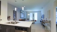 This single-floor space showcases a contemporary open-concept design that effortlessly blends the kitchen and living areas into one cohesive and functional environment. The kitchen features a clean white countertop island with an integrated sink and stainless steel dishwasher, complemented by bold pendant lighting that adds a modern, industrial touch to the space. Soft blue-gray walls create a calm and inviting atmosphere throughout, while neutral-toned carpeting in the living area adds warmth underfoot. The living space is generously proportioned, offering ample room for comfortable seating arrangements, including a plush sofa and a distinctive hanging egg chair that adds a playful design element. Large windows allow natural light to pour in, brightening the interior and providing views of the outdoors. A flat-screen television is mounted on a media console along one wall, while an eye-catching large-scale framed artwork and a surf-culture typography poster add personality and character to the décor. Recessed lighting throughout further enhances the bright and airy ambiance of the space, making it both visually appealing and highly livable.