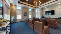 Located at 161 Grande Oaks Dr in Charleston, SC, this single-floor screening and media room is thoughtfully designed for a premium viewing experience. The space features rows of plush, cognac-toned leather lounge chairs arranged in a theater-style configuration, providing comfortable and unobstructed sightlines to a large wall-mounted flat-screen television. The elevated tray ceiling, finished in a rich dark wood tone with decorative molding, adds architectural sophistication and a sense of height to the room. Soft, neutral wall panels with classic wainscoting detailing line the perimeter, complemented by an abundance of windows fitted with roller shades that allow for natural light control. The flooring throughout is a deep blue-gray carpet that enhances the room's cinematic atmosphere. Along one wall, a sleek built-in credenza with wood-grain cabinetry and a dark countertop provides ample storage and surface space, accompanied by an under-counter beverage refrigerator — ideal for entertaining. A contemporary multi-bulb chandelier adds a stylish focal point near the entrance. Film-themed artwork and tasteful decorative accents complete the curated aesthetic, making this space a well-appointed venue for screenings, gatherings, or media events.