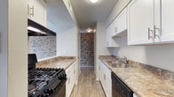Located in Laurel, this single-floor residence offers a thoughtfully designed layout with functional and stylish interiors. The galley-style kitchen is a standout feature, showcasing crisp white cabinetry with brushed nickel hardware, granite-look countertops, and a full suite of black appliances including a gas range, refrigerator, and dishwasher. A stainless steel sink with a functional faucet adds a practical touch, while the textured ceiling and wood-look flooring lend warmth and character to the space. A distinctive brick accent wall runs along the kitchen corridor, adding a rustic, industrial charm that contrasts beautifully with the clean white cabinetry. The kitchen opens into a hallway that connects to the rest of the home. The living area features plush carpeting and a window that invites natural light, creating a bright and welcoming atmosphere. Neutral wall tones throughout the space provide a versatile backdrop for a variety of interior styles. Overall, the residence combines practical design with inviting finishes, making it a comfortable and functional living space in Laurel.