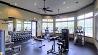 This fitness center offers a comprehensive workout environment designed to accommodate a range of exercise routines. The space features dark-trimmed walls with a warm neutral color palette, lending it a polished, club-like atmosphere. Two ceiling fans are suspended overhead, complementing the recessed lighting to maintain comfort during workouts. Large windows along one wall flood the room with natural light while offering views of surrounding greenery, creating an open and energizing ambiance. A glass door provides additional access and reinforces the connection to the outdoors. The flooring consists of dark, durable material suited for heavy equipment use. Strength training is well-supported with a multi-station weight machine, a dedicated free weights rack stocked with dumbbells, and a flat workout bench. Cardio equipment includes elliptical trainers, treadmills, and a stair climber, offering diverse aerobic options. A large mirror along one wall expands the sense of space and allows users to monitor their form. The overall layout is thoughtfully organized, ensuring adequate spacing between machines for safe and efficient use throughout the room.
