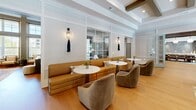 Located at 382 Red Bud Cir in Hendersonville, NC, this thoughtfully designed single-floor community space offers a comfortable and elegant setting across its living and dining areas. The living room showcases a refined aesthetic with coffered ceilings, white wainscoting panel walls, and warm hardwood flooring throughout. A sleek linear fireplace serves as the focal point, flanked by large windows that invite natural light and views of the surrounding greenery. A wall-mounted TV above the fireplace and plush, neutral-toned sofas complete the inviting atmosphere. An adjacent lounge area features a long caramel leather banquette paired with curved velvet swivel chairs and marble-topped pedestal tables, accented by elegant tassel wall sconces. The dining room is defined by sage-green shiplap walls, a substantial natural wood dining table accommodating multiple guests, and a dramatic cluster of globe pendant lights overhead. Built-in shelving and a textured tile accent wall add depth and character to the room. A secondary dining nook with richly upholstered chairs and moody wallpaper provides an intimate setting. Glass double doors open directly to an outdoor pool area, seamlessly blending the interior with the exterior landscape.