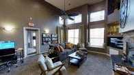 This single-floor community lounge is designed to impress with its dramatic, soaring double-height ceilings that create an immediate sense of openness and grandeur. A large ceiling fan anchors the central seating area, complementing the warmth and airflow of the well-appointed space.

The lounge is bathed in natural light, courtesy of tall, multi-pane windows dressed with flowing neutral drapes and adjustable blinds. A built-in window seat offers a cozy nook perfect for relaxing or reading. The walls are painted in a rich, sophisticated charcoal-gray tone that lends the space a modern and refined ambiance.

Furnishings are carefully curated for both style and comfort. A plush, dark gray sofa adorned with burnt orange and patterned accent pillows serves as the focal point of the main seating arrangement, flanked by tasteful armchairs — including a mid-century modern wood-framed accent chair. A sleek, two-tier coffee table ties the seating area together.

A wall-mounted literature rack near the entrance provides a practical touch, keeping reading materials and community resources neatly organized and accessible. The patterned carpeting adds texture and warmth underfoot, contributing to the inviting, residential feel of this thoughtfully designed communal gathering space.