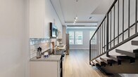 Located on E Adams Street in the heart of downtown Jacksonville, Florida, this two-story, one-bedroom, two-bathroom residence offers a well-organized layout spread across two evenly sized floors. The ground floor features hardwood-style flooring throughout, setting a warm and modern tone. A dedicated laundry room houses a stacked washer and dryer unit, providing everyday convenience. Two full bathrooms are finished with granite-topped white vanities, sleek rectangular mirrors, and brushed nickel fixtures. One bathroom includes a tiled soaking tub and shower combination, while the other serves as an en-suite accessible from the bedroom. The upper floor is home to the generously sized primary bedroom, which stands out with its exposed concrete ceiling — a striking industrial design element — paired with a ceiling fan and plush wall-to-wall carpeting. Multiple windows flood the room with natural light and offer views of the surrounding urban neighborhood. A dedicated walk-in closet fitted with wire shelving and carpeted flooring provides ample storage. The staircase connecting the two floors features a dark handrail that contrasts neatly with the light-toned walls and carpet. Throughout the residence, a neutral gray and white palette creates a clean, contemporary ambiance well-suited to urban living.