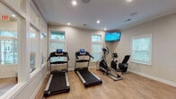 This fitness center in Gulf Shores, Alabama, offers a well-appointed, single-level exercise space designed to support a well-rounded workout routine. The room features warm-toned hardwood-style flooring throughout, neutral gray walls, and crown molding that lend a polished, residential feel to the space. Recessed lighting is complemented by multiple windows fitted with white plantation blinds, allowing natural light to filter in while maintaining privacy.

The cardio area is equipped with two treadmills and an elliptical trainer, each featuring integrated digital displays, as well as a recumbent stationary bike. A wall-mounted flat-screen television is positioned at the front of the cardio section, providing entertainment during workouts. The strength training area includes a full rack of dumbbells spanning a wide range of weights, a wall-mounted exercise mat, and an adjustable workout bench, offering ample options for resistance training.

A glass partition wall with interior windows separates the fitness center from an adjacent lounge area, adding architectural interest and visual openness. A dedicated entry door provides direct access to the exterior. The overall ambiance is clean, bright, and functional, making this fitness center a practical and inviting amenity for users seeking a convenient place to exercise.
