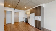 This thoughtfully designed one-bedroom, one-bathroom unit offers a well-organized single-floor layout that maximizes every square foot. The open-concept living and kitchen area immediately draws attention to the striking exposed concrete ceiling, which lends an industrial-chic character throughout the space. Warm wood-look flooring runs seamlessly through the main living area, complementing the kitchen's dark wood-tone cabinetry.

The kitchen is efficiently appointed with a full suite of white appliances, including a refrigerator, electric range, dishwasher, and built-in microwave. A stainless steel range hood, open shelving, and ample cabinet storage complete this functional cooking space. Recessed lighting in the kitchen area adds a polished touch against the raw concrete ceiling.

The living area benefits from a large window that invites natural light and provides views of the surrounding urban streetscape. The separate bedroom features soft carpeting underfoot and its own window with an exterior view, creating a quiet, comfortable retreat.

The bathroom is clean and functional, finished in crisp white with a full soaking tub and shower combination. The overall aesthetic of the unit balances industrial elements with modern, minimalist finishes, resulting in a cohesive and practical living space.