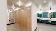 This single-floor space blends functional design with striking aesthetic choices throughout. The restroom area is a standout feature, finished with bold, glossy teal subway tiles that wrap the walls from floor to mid-height, creating a vivid and energetic atmosphere. A continuous white countertop with undermount sinks and matte black fixtures runs the length of the vanity wall, complemented by arched mirrors and globe pendant lights suspended from warm wood-finished ceiling mounts. Polished concrete floors ground the space with an industrial edge, while stainless steel stall partitions add a sleek, utilitarian counterpoint. Adjacent to the restrooms, a generous locker area provides ample storage through floor-to-ceiling natural wood-paneled lockers, offering a warm contrast to the cool tones nearby. Globe flush-mount ceiling fixtures provide clean, even illumination throughout this zone. A separate lounge area offers a more intimate setting, featuring a tufted leather sofa, layered accent lighting, botanical-patterned wallcovering in deep navy tones, and curated wall art. Large windows in the lounge frame views of an outdoor courtyard with slatted wood fencing and surrounding urban architecture, allowing natural light to fill the space and connect the interior to its surroundings.