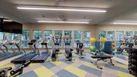 This fitness center is thoughtfully designed to support a wide range of workout routines in a bright, welcoming environment. The space is divided into distinct functional zones, seamlessly connected by a bold, color-blocked carpet in shades of blue, yellow, and gray that adds a lively visual character to the room.

The cardio zone is positioned along a wall lined with multiple double-hung windows that invite abundant natural light and offer views of surrounding greenery. Treadmills, elliptical trainers, and upright and recumbent stationary bikes are arranged in a row, each equipped with digital displays. A row of mirrors runs along the adjacent wall, visually expanding the space and allowing users to monitor their form.

The strength training area occupies a separate section, distinguished by its blue-painted walls and a large wall-mounted flat-screen television. A multi-station cable machine serves as the centerpiece, complemented by a dumbbell rack stocked with a full range of weights, a weight bench, and a compact beverage refrigerator nearby. An assortment of medicine balls, stability balls, foam rollers, and resistance accessories are neatly stored on a dedicated rack. Overhead linear LED lighting and ceiling fans ensure the space remains well-lit and comfortable throughout.