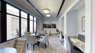 This elegantly designed single-floor lounge space offers a refined and functional environment suited for a variety of uses. The room is finished with soft blue-grey walls, crisp white crown molding, and a patterned herringbone-style carpet that adds visual texture underfoot. Large floor-to-ceiling windows along one wall flood the interior with natural light, offering views of a lush outdoor courtyard and creating an open, airy atmosphere.

The seating area features a curated arrangement of upholstered armchairs and accent chairs in neutral tones of beige and grey, gathered around dark-stained circular tables. This lounge zone is well-suited for casual meetings or social gatherings. Overhead, elegant dome pendant light fixtures complement the recessed ceiling lighting, contributing to the space's polished aesthetic.

Along the opposite wall, a series of built-in alcoves house individual workstations with countertop surfaces and task chairs, providing semi-private areas for focused work. A triptych of abstract framed artwork adorns the wall above, adding a contemporary artistic touch. The overall ambiance is professional yet comfortable, balancing collaborative and individual needs seamlessly.