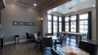 This sophisticated single-floor community lounge is designed for both relaxation and socializing. The space is anchored by a sleek billiards table positioned near a wall of tall, dramatic windows dressed with floor-length dark curtains, allowing generous natural light to fill the room while offering views of the surrounding building exterior. The hardwood-style flooring runs throughout, lending warmth and cohesion to the overall aesthetic.

The layout is thoughtfully divided into distinct seating zones. Dark wood dining tables paired with tufted upholstered chairs create intimate gathering areas ideal for casual meetings or game nights. A pendant light fixture adds a refined focal point above the billiards area, while recessed ceiling lighting and wall sconces provide layered illumination throughout. Crown molding along the ceiling adds a subtle architectural elegance to the space.

The walls are painted in a cool, neutral gray tone and accented with a curated gallery of framed artwork, enhancing the contemporary yet inviting ambiance. The overall design strikes a balance between upscale style and comfortable functionality, making this clubroom a welcoming destination for recreation and community engagement.