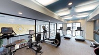 This contemporary fitness center is thoughtfully designed to provide an energizing and comfortable workout environment. The space is defined by its striking teal-painted ceiling accents set within a coffered ceiling framework, complemented by recessed lighting and track fixtures that ensure the room is bright and welcoming throughout.

The cardio equipment offering is extensive, featuring multiple treadmills, elliptical cross-trainers, and recumbent cycles from Life Fitness — all arranged to maximize space and usability. A wall-mounted flat-screen television is positioned for easy viewing during workouts, adding an entertainment element to the fitness experience. Patterned carpet flooring in a neutral tone ties the space together while providing comfort underfoot.

Large floor-to-ceiling windows and glass partition walls flood the fitness center with abundant natural light and offer open views of the surrounding building exterior. The glass partitions also provide a visual connection to an adjacent lounge area, which features warm yellow walls, decorative tile accents, a communal table with seating, and a distinctive starburst chandelier — creating a lively, social atmosphere just steps away.

Lush potted plants and bold geometric wallcovering accents add a decorative touch, while the overall design reflects a modern, upscale aesthetic that balances functionality with style.