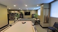 This thoughtfully designed single-floor space functions as a versatile recreation and lounge area, combining entertainment and relaxation in a cohesive, welcoming environment. At the heart of the room sits a full-sized billiards table, serving as the centerpiece for social gatherings and leisure activity. Surrounding it, a comfortable seating arrangement faces a wall-mounted flat-screen television, creating an inviting media lounge area ideal for unwinding or watching events together. The room is lined with tall, fully stocked bookshelves, adding both visual interest and a warm, intellectual character to the space. Potted greenery is strategically placed throughout, introducing natural elements that soften the room's neutral, earthy color palette of warm taupes and grays. Recessed ceiling lighting provides even, ambient illumination across the entire space, ensuring a well-lit atmosphere at any time of day. A window with blinds allows for natural light to filter in while maintaining privacy. The flooring features low-pile carpet that complements the overall tonal scheme. A mounted cue rack on the wall near the billiards area adds a practical and organized finishing touch. Overall, the space strikes a balance between functionality and comfort.