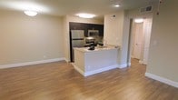 This well-appointed single-floor space showcases a thoughtfully designed open-concept layout that seamlessly connects the kitchen and living areas. The kitchen is a standout feature, equipped with stainless steel appliances including a full-size refrigerator, microwave, and range. Rich dark cabinetry provides ample storage, while granite countertops lend a polished, upscale feel. A peninsula-style counter extends into the living space, offering additional prep area and casual seating opportunities. The open layout creates a natural flow between the kitchen and the adjoining living area, making the space feel expansive and inviting. Warm hardwood-style flooring runs continuously throughout, adding texture and cohesion to the overall design. Neutral-toned walls and flush ceiling lighting contribute to a clean, bright interior atmosphere. A hallway visible in the background leads to additional areas of the unit, suggesting a practical and well-organized floor plan. Louvered closet doors hint at generous storage solutions. The overall aesthetic balances contemporary finishes with functional design, making this a comfortable and visually appealing living environment.
