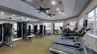 This fitness center offers a thoughtfully designed single-floor workout space that balances functionality with a welcoming atmosphere. The room features an angled, tray-style ceiling equipped with multiple ceiling fans, ensuring comfortable airflow throughout even the most intense workout sessions. Recessed lighting supplements the abundant natural light that streams in through a series of large, multi-pane windows lining the walls, providing views of the surrounding landscape and creating an open, airy environment. The space is outfitted with a comprehensive selection of Life Fitness and other brand-name equipment, including treadmills, an elliptical machine, and a variety of cable and selectorized strength training machines suited for both upper and lower body workouts. Full-length mirrors along one wall visually expand the room and allow users to monitor their form during exercise. The neutral-toned carpeted flooring provides a soft underfoot surface throughout the space. The overall layout is spacious and well-organized, allowing users to move freely between cardio and strength stations. The combination of natural light, ceiling fans, quality equipment, and thoughtful design makes this fitness center a practical and pleasant environment for daily exercise.