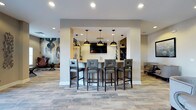 This single-floor space presents a well-appointed lounge and bar area designed for socializing and relaxation. At the heart of the space is a built-in bar counter framed by a structural archway, equipped with a light-toned stone or quartz countertop and surrounded by five upholstered bar-height chairs in a sleek gray finish. Four matte black dome pendant lights hang above the counter, adding a modern industrial touch that complements the recessed lighting throughout the ceiling.

Behind the bar, wall-mounted cabinetry and a flat-screen television create a functional and inviting focal point. The open-concept layout extends into a broader lounge area, where luxury vinyl plank flooring in a stone-look finish ties the space together with a neutral, contemporary aesthetic. The neutral beige and white wall tones provide a clean backdrop, while a large abstract wall mural on the left side introduces a bold artistic element. Additional seating, including upholstered lounge chairs, is visible in the surrounding areas, reinforcing the communal and comfortable character of the space. The overall ambiance is modern, refined, and well-suited for casual gatherings.
