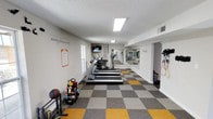 This single-floor fitness center is designed to offer a comprehensive workout experience in a clean and energizing environment. The space is anchored by a row of treadmills and cardio machines aligned along one wall, providing a structured area for aerobic exercise. A full-length mirror spans the far wall, enhancing the sense of depth and allowing users to monitor their form during workouts.

The flooring features a distinctive checkered pattern in shades of grey, white, and yellow, adding a dynamic visual element to the space. A free weight station near the entrance houses a variety of dumbbells and kettlebells neatly arranged on a rack, catering to strength training needs. Resistance bands and additional accessories are also visible, offering versatile workout options.

Motivational wall decals and quotes, including phrases such as "Activity fuels more activity," contribute to an upbeat and encouraging atmosphere. A wall-mounted flat-screen TV is positioned for easy viewing during cardio sessions. Natural light filters in through a window on one side, complemented by recessed fluorescent ceiling fixtures that ensure the space is well-lit throughout. The textured white ceiling and neutral-toned walls keep the overall aesthetic clean and focused on functionality.