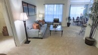 This single-floor residence offers a well-appointed living space designed for both comfort and style. The living area serves as the heart of the home, featuring plush wall-to-wall carpeting that adds warmth underfoot. Large windows dressed with sheer curtains flood the room with abundant natural light, creating an airy and open ambiance throughout the day. The space is thoughtfully furnished with a generous sofa accented by warm copper and neutral throw pillows, complemented by an elegant glass-topped coffee table with ornate metal legs. A classic floor lamp anchors one corner of the room, providing additional warm lighting. Bold abstract artwork adorns the walls, adding a modern artistic touch to the neutral color palette. Adjacent to the main seating area, a dining space is visible, furnished with sleek chairs and a potted plant that introduces a refreshing natural element. The overall design balances contemporary accents with traditional furnishings, resulting in a cohesive and welcoming interior. The open layout allows for flexible use of the space, making it well-suited for both everyday living and entertaining guests.