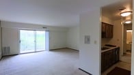 This single-floor space features an open-concept living area finished with wall-to-wall carpeting and neutral white walls that create a bright, clean atmosphere. The room is anchored by a large sliding glass door fitted with vertical blinds, which opens to a private balcony overlooking a lush, green hillside — a standout feature that brings natural light deep into the interior. Ventilation is provided through a ceiling duct, and standard electrical outlets are distributed along the baseboards. Adjacent to the main living area, a partial wall separates the kitchen, which is visible from the living space and features dark cabinetry and a light-toned countertop, suggesting a functional layout with convenient access between the two areas. The overall design is straightforward and practical, with an emphasis on open space and connectivity to the outdoors. The neutral palette throughout allows for flexible furnishing and décor options, while the natural light provided by the oversized sliding door gives the space an airy, welcoming character.