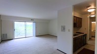This single-floor space features an open-concept living area finished with wall-to-wall carpeting and neutral white walls that create a bright, clean atmosphere. The room is anchored by a large sliding glass door fitted with vertical blinds, which opens to a private balcony overlooking a lush, green hillside — a standout feature that brings natural light deep into the interior. Ventilation is provided through a ceiling duct, and standard electrical outlets are distributed along the baseboards. Adjacent to the main living area, a partial wall separates the kitchen, which is visible from the living space and features dark cabinetry and a light-toned countertop, suggesting a functional layout with convenient access between the two areas. The overall design is straightforward and practical, with an emphasis on open space and connectivity to the outdoors. The neutral palette throughout allows for flexible furnishing and décor options, while the natural light provided by the oversized sliding door gives the space an airy, welcoming character.