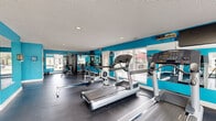 This fitness center is a thoughtfully designed, single-story facility that offers a comprehensive workout environment for users of all fitness levels. The interior is immediately distinguished by its bold teal accent walls, which create an energetic and motivating atmosphere complemented by a neutral textured ceiling and durable dark rubber flooring throughout.

The cardio zone is well-stocked with multiple treadmills, rowing machines, an elliptical trainer, and a stationary bike, providing ample options for cardiovascular exercise. A dedicated strength training area features a cable and weight machine setup, rounding out the facility's full-body workout capabilities. Large wall-mounted mirrors expand the sense of space and allow users to monitor their form during exercise.

Natural light pours in through multiple large windows fitted with horizontal blinds, offering pleasant views of the surrounding greenery and keeping the space feeling open and airy. Recessed lighting in the ceiling ensures consistent illumination throughout all hours of operation.

The facility is accessed through a private entrance door, and the exterior features a classic red brick facade with a spacious adjoining wood-composite deck shaded by mature trees — providing a welcoming outdoor area just steps from the workout floor. The overall design is clean, functional, and purpose-built to support an active lifestyle.