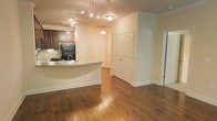 This single-floor residence offers an open-concept layout that seamlessly connects the kitchen and living areas, creating a cohesive and functional living environment. Rich hardwood flooring runs throughout the main space, lending warmth and a sense of continuity to the interior.

The kitchen is positioned along one side of the open floor plan and features dark wood cabinetry, a black refrigerator, and a peninsula with a light-toned countertop that doubles as a breakfast bar. A pendant light hangs above the peninsula, while a track lighting system provides additional illumination over the kitchen workspace.

The living area is generously proportioned, benefiting from the open layout and warm, neutral wall tones that create an inviting ambiance. Crown molding runs along the ceiling line, adding a refined architectural detail throughout the space. A flush-mount ceiling light further brightens the main living zone.

Several interior doors are visible along the back wall, indicating access to additional rooms or storage, including what appears to be a closet. The overall design balances classic detailing with practical, everyday functionality.