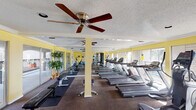 Located at 2400 Westheimer Rd in Houston's vibrant Montrose neighborhood, this fitness center offers a comprehensive and well-organized workout facility on a single floor. The space is defined by its bright yellow accent walls, textured ceilings, and large windows that line the perimeter, flooding the room with natural light and offering views of the surrounding outdoor areas. Ceiling fans are installed throughout, ensuring comfortable air circulation during workouts. The main exercise area is generously proportioned and features a row of treadmills as the centerpiece, complemented by an elliptical trainer, a stationary bike, and a multi-station cable weight machine. A dedicated free weights section includes a full dumbbell rack, weight benches, and additional strength training equipment. Floor-to-ceiling mirrors are strategically placed along one wall, visually expanding the space and allowing users to monitor their form. A secondary area near the entrance provides additional utility space, housing stability balls and other accessories. The fitness center is accessible via French doors that open to an outdoor walkway, creating a seamless connection to the exterior. Carpet flooring covers the main workout area, and the overall ambiance is clean, functional, and inviting—well-suited for a variety of fitness routines.