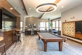 Located at 2071 Powers Ferry Rd SE in Marietta, Georgia, this single-story community clubhouse delivers an impressive blend of rustic and contemporary design elements. The space is anchored by soaring vaulted ceilings adorned with exposed dark wood beams and dramatic cross-beam accents, creating an open, airy atmosphere throughout.

The main lounge area is a standout feature, centered around a floor-to-ceiling stacked stone fireplace with a rich wood mantel — a commanding focal point that adds warmth and character to the room. Comfortable seating, including a large leather sectional sofa and neutral-toned accent chairs arranged around a round coffee table, invites relaxed gathering and socializing.

Wide-plank wood-look flooring runs throughout the space, complementing the warm wood tones of built-in cabinetry along one wall, which provides ample storage and a polished, finished look. A dedicated bar-height dining area with bar stools offers a casual spot for conversation or refreshments, while additional seating areas with sleek modern chairs are positioned near large windows that draw in natural light and offer views of the surrounding greenery.

Curated wall art and thoughtful furnishings round out the interior, lending a refined yet approachable aesthetic. The overall ambiance is welcoming and versatile, well-suited for community events, casual lounging, or everyday resident use.