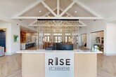 Located at 100 Inspire Way in St. Augustine, FL, this thoughtfully designed single-floor community clubhouse offers an expansive, open layout that combines modern aesthetics with functional versatility. The architectural design is immediately striking, anchored by dramatic vaulted ceilings with exposed white-painted timber trusses that draw the eye upward and flood the interior with an airy, light-filled atmosphere. Large clerestory windows and glass entry doors allow abundant natural light to pour throughout the space.

Light-toned herringbone hardwood flooring runs continuously across the entire floor, lending warmth and visual cohesion to each area. At the entrance, a curved reception desk with fluted wood detailing and a branded accent wall sets a welcoming, professional tone. The spacious living room features a comfortable lounge seating arrangement, a shuffleboard table, a wall-mounted television, and built-in shelving with warm wood finishes and tile backsplash accents. A sleek marble-topped bar counter with blue upholstered stools provides a casual gathering spot, while a billiards table adds to the entertainment offerings.

A vertical slat partition elegantly defines zones within the open floor plan without sacrificing sightlines. The office area includes glass-enclosed private work booths, each furnished with a built-in desk, pendant lighting, and a natural-light-filled window — ideal for focused individual use. Throughout, curated artwork and contemporary furnishings create a polished, inviting ambiance.