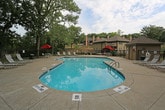 This inviting outdoor amenity space centers around a generously sized, kidney-shaped swimming pool with crystal-clear turquoise water. The pool deck offers ample room for relaxation, lined with white lounge chairs and shaded by red market umbrellas positioned at various seating areas — ideal for enjoying warm days in comfort. The entire pool area is enclosed by a sturdy black iron fence, providing both security and an open, airy feel that allows unobstructed views of the mature, towering trees that surround the property. A covered patio area adjacent to the pool features wood-beam support posts and a secure gated entry, with a concrete pad that serves as a transitional space between the building and pool deck. The building exterior is finished in light horizontal lap siding with a sage green entry door accented by sidelight windows with blinds, adding a welcoming touch to the overall aesthetic. Posted pool rules and designated hours reflect a well-managed facility. The lush greenery and natural canopy of trees provide a serene, park-like atmosphere throughout the space, making it a standout outdoor retreat within the community.