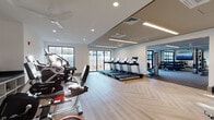 Located at 105 Corporate Park Dr in West Harrison, NY, this contemporary fitness center is thoughtfully designed to support a comprehensive workout experience across a single well-organized floor. The main cardio area features a row of treadmills and elliptical machines, all positioned along large floor-to-ceiling windows that flood the space with natural light and provide open outdoor views. The light-toned herringbone hardwood flooring adds a refined, modern aesthetic throughout the primary exercise area. A dedicated functional training zone houses a Life Fitness Synrgy360 multi-station system, accompanied by medicine balls, TRX suspension trainers, and additional accessories, all set on durable rubberized flooring suited for dynamic movements. Adjacent to this, a strength training room features a full rack of dumbbells, weight benches, and mirrored walls that visually expand the space. A separate open area with resilient flooring and expansive glass garage-style doors offers versatility for group or individual training. Recessed and linear LED lighting throughout ensures consistent illumination, while ceiling fans and integrated HVAC systems maintain comfort. A wall-mounted flat-screen display in one section provides an engaging visual focal point. The overall design balances functionality with a clean, bright, and professional atmosphere.