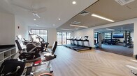 Located at 105 Corporate Park Dr in West Harrison, NY, this contemporary fitness center is thoughtfully designed to support a comprehensive workout experience across a single well-organized floor. The main cardio area features a row of treadmills and elliptical machines, all positioned along large floor-to-ceiling windows that flood the space with natural light and provide open outdoor views. The light-toned herringbone hardwood flooring adds a refined, modern aesthetic throughout the primary exercise area. A dedicated functional training zone houses a Life Fitness Synrgy360 multi-station system, accompanied by medicine balls, TRX suspension trainers, and additional accessories, all set on durable rubberized flooring suited for dynamic movements. Adjacent to this, a strength training room features a full rack of dumbbells, weight benches, and mirrored walls that visually expand the space. A separate open area with resilient flooring and expansive glass garage-style doors offers versatility for group or individual training. Recessed and linear LED lighting throughout ensures consistent illumination, while ceiling fans and integrated HVAC systems maintain comfort. A wall-mounted flat-screen display in one section provides an engaging visual focal point. The overall design balances functionality with a clean, bright, and professional atmosphere.