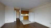 This single-floor unit presents an open-concept layout that integrates the living area and kitchen into one cohesive space. The kitchen is defined by warm medium-toned wood cabinetry overhead and below the counter, providing ample storage. It is equipped with a white gas range, a double-basin sink, and a full-size white refrigerator, all set atop light ceramic tile flooring that visually distinguishes the cooking zone from the rest of the unit. The surrounding living area features dark hardwood flooring, creating a natural contrast between the two spaces. White walls and a white ceiling reflect light throughout, keeping the interior feeling open and clean. A hallway visible from the main space leads to the unit's entry door, which is fitted with a wall-mounted light fixture. The overall layout is practical and straightforward, with a neutral color palette that offers flexibility for personalization. The space is compact yet efficiently designed, making good use of the available floor plan to accommodate everyday living needs within a single level.