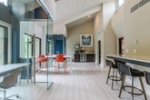 Located at 3000 Cumberland Club Dr in Atlanta, Georgia, this single-floor amenity space combines contemporary design with highly functional work and meeting environments. The layout features soaring vaulted ceilings with white-paneled soffits in the main open area, light wood-look flooring, and an abundance of natural light streaming through large black-framed windows that frame views of mature, verdant tree canopy. A built-in bar counter with bar-stool seating provides a casual workspace or gathering spot, complemented by a credenza with beverage storage and a wet bar along the back wall. Floor-to-ceiling glass partitions define two distinct enclosed meeting rooms, maintaining visual openness while offering privacy. One room features a warm wood conference table with blue upholstered chairs and bold abstract artwork, while the other accommodates a round table with mustard-yellow seating — ideal for collaborative sessions. A private lounge nook with plush dark seating and a bright red accent table sits beside a large window overlooking the greenery. An additional semi-private workstation enclosed in glass offers a focused individual workspace. The entry corridor features patterned carpet tile, a decorative console alcove, and access to a private room. Throughout, a curated palette of bold accent colors, contemporary furnishings, and thoughtful artwork creates a vibrant yet professional atmosphere.