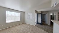 This single-floor residence offers a thoughtfully designed layout with a clean, neutral color palette and a blend of hard-surface and carpeted flooring throughout. The living area is generously sized, featuring plush wall-to-wall carpet, soft gray walls, and multiple windows dressed with horizontal blinds that invite an abundance of natural light. A bold navy entry door with a sidelight window adds a striking focal point to the main living space.

The kitchen is well-equipped with light taupe cabinetry, a breakfast bar peninsula, light countertops, and stainless steel appliances including a refrigerator and microwave. Wood-look vinyl plank flooring flows through the kitchen and dining areas, creating a cohesive transition between spaces.

The bedroom continues the warm, carpeted aesthetic and is fitted with a ceiling fan with light fixture, providing both comfort and functionality. A window with blinds allows for natural light while maintaining privacy.

The bathroom features a single-sink vanity with light wood cabinetry, a large mirror, modern three-bulb vanity lighting, and wood-look flooring. The layout is efficient and well-organized. Overall, the space presents a practical, move-in-ready environment with consistent finishes and a comfortable, understated ambiance throughout.
