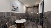 Located at 611 S Street in Baltimore, MD, this single-level dog wash facility is thoughtfully designed to provide a functional yet polished environment for pet grooming. The space features a cohesive aesthetic defined by large-format dark charcoal stone-look tiles that line the walls and floors throughout, lending a sleek, modern character to every area.

The wash station area serves as the centerpiece of the facility, equipped with multiple handheld shower heads mounted on adjustable rails along fully tiled walls, allowing for thorough and flexible bathing. A low-profile stainless steel step platform sits within the wash zone, accommodating dogs of varying sizes. The wet area transitions seamlessly into a grooming and drying zone, where a built-in counter with a stainless steel sink provides a dedicated workspace. Wall-mounted outlets and hooks support grooming tools, and a professional-grade dryer is conveniently stationed nearby.

Wood-look plank flooring runs through the central area of the space, adding warmth against the cool tile surroundings. A round marble-top table with a rustic wood base sits in the open area, providing a welcoming focal point. Recessed ceiling lighting ensures the space is well-illuminated throughout. Playful dog-themed artwork displayed on the upper white walls adds personality and a lighthearted ambiance to this well-appointed pet care facility.