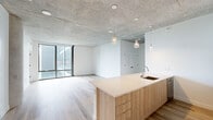 This thoughtfully designed one-bedroom, one-bathroom residence offers a compelling blend of industrial and contemporary aesthetics across a well-proportioned single floor. The open-concept kitchen and living area serves as the heart of the space, anchored by a large island with a warm wood-toned base and a light quartz countertop, an undermount sink, and pendant lighting suspended from a striking exposed concrete ceiling. The kitchen is outfitted with white upper cabinetry, a subway tile backsplash, stainless steel appliances including a refrigerator, built-in microwave, and range. Light hardwood-style flooring runs throughout, complementing the clean white walls and raw concrete overhead. Floor-to-ceiling windows in both the living area and bedroom flood the space with natural light and frame elevated outdoor views of the surrounding cityscape and greenery. The bedroom offers a comfortable footprint with direct access to its own floor-to-ceiling window wall, along with an exposed ductwork detail that reinforces the industrial character. The bathroom features a wood-vanity with a white countertop, a large wall mirror, and a full tub-shower combination wrapped in marble-style tile. Two hallways provide practical connectivity throughout the layout.